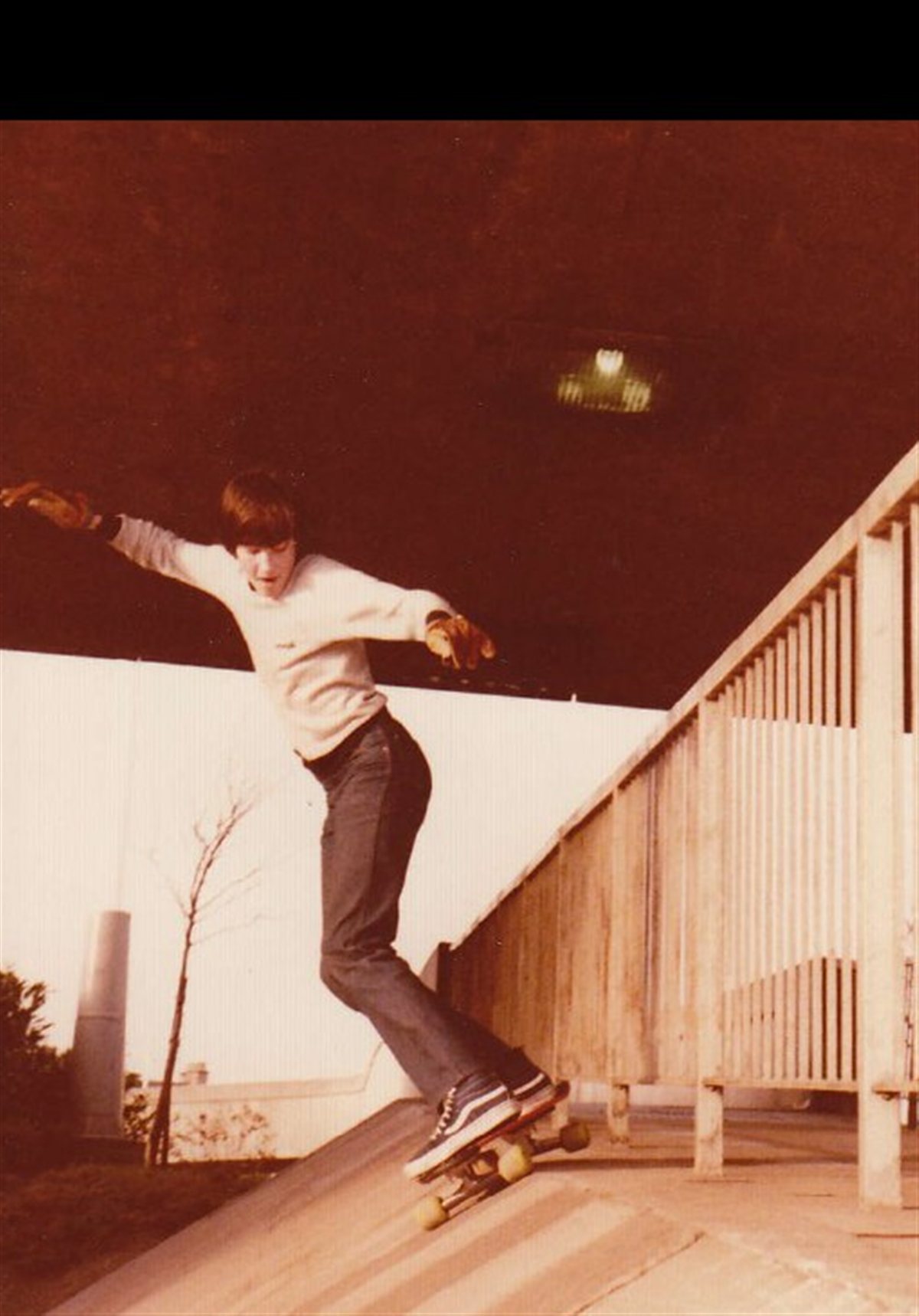 A young skater in late-1970s clothing &mdash; sweatshirt, jeans, Vans &mdash; riding a 45-degree paved bank under a road overpass, with a wooden side-railing running up the bank. From Mike O'Brien's personal collection in The Bloody Kids' Book.