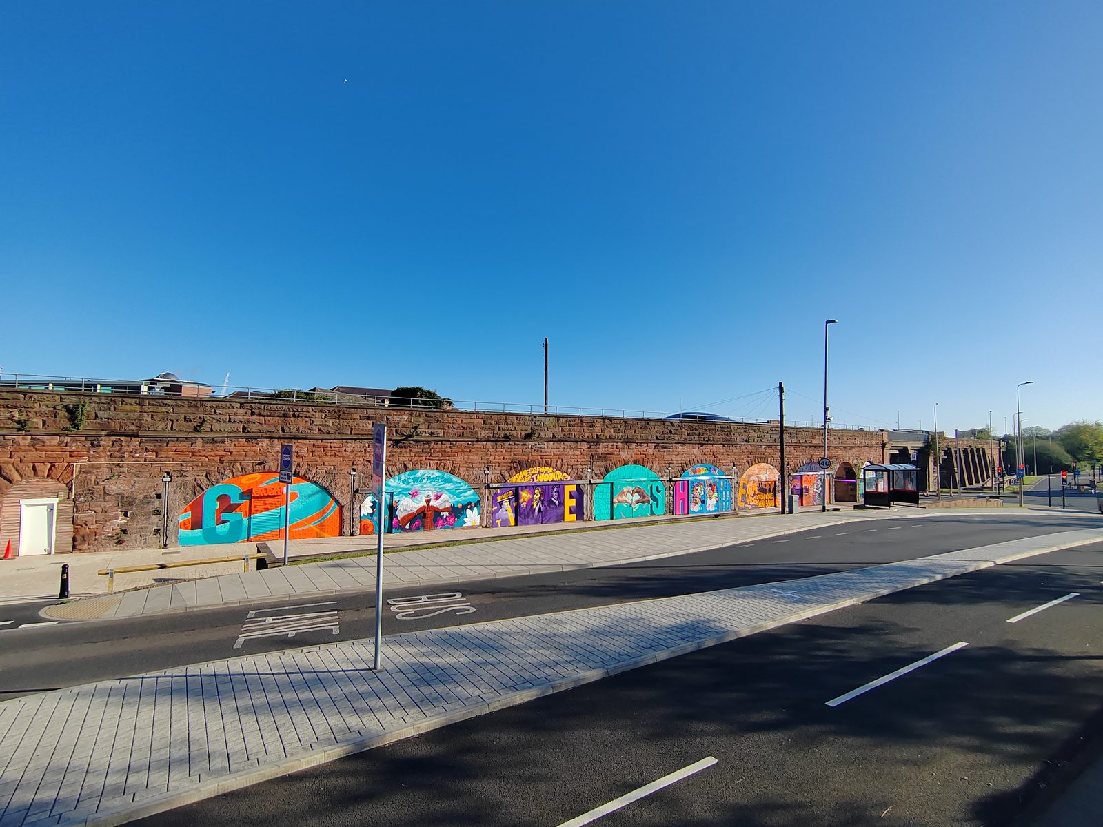 Wide panoramic view of the Askew Road site from across the road, showing the full run of seven railway arches with colourful murals, the bus lane in the foreground, and the urban context.