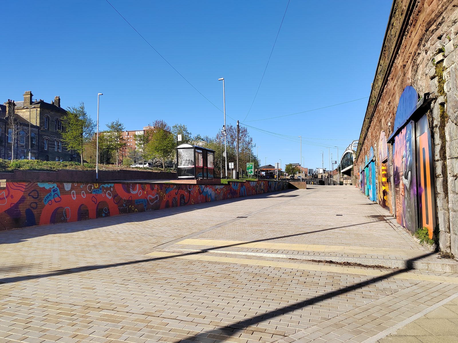 Long view from the east end of the site looking back along the arches, showing the road, bus shelter, residential context on the hill above, and pedestrian access.